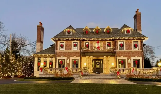 Large brick colonial with a lit wreath in every window and garland along the porch