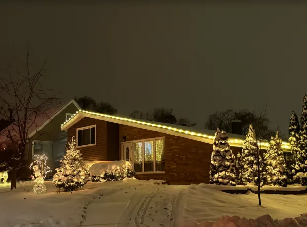 Snow-covered ranch with warm white roofline and wrapped evergreens