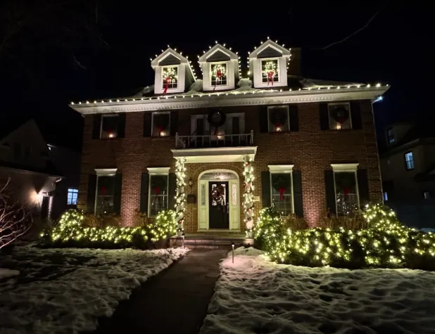 Brick colonial with lit wreaths on the dormers and windows over a snowy lawn