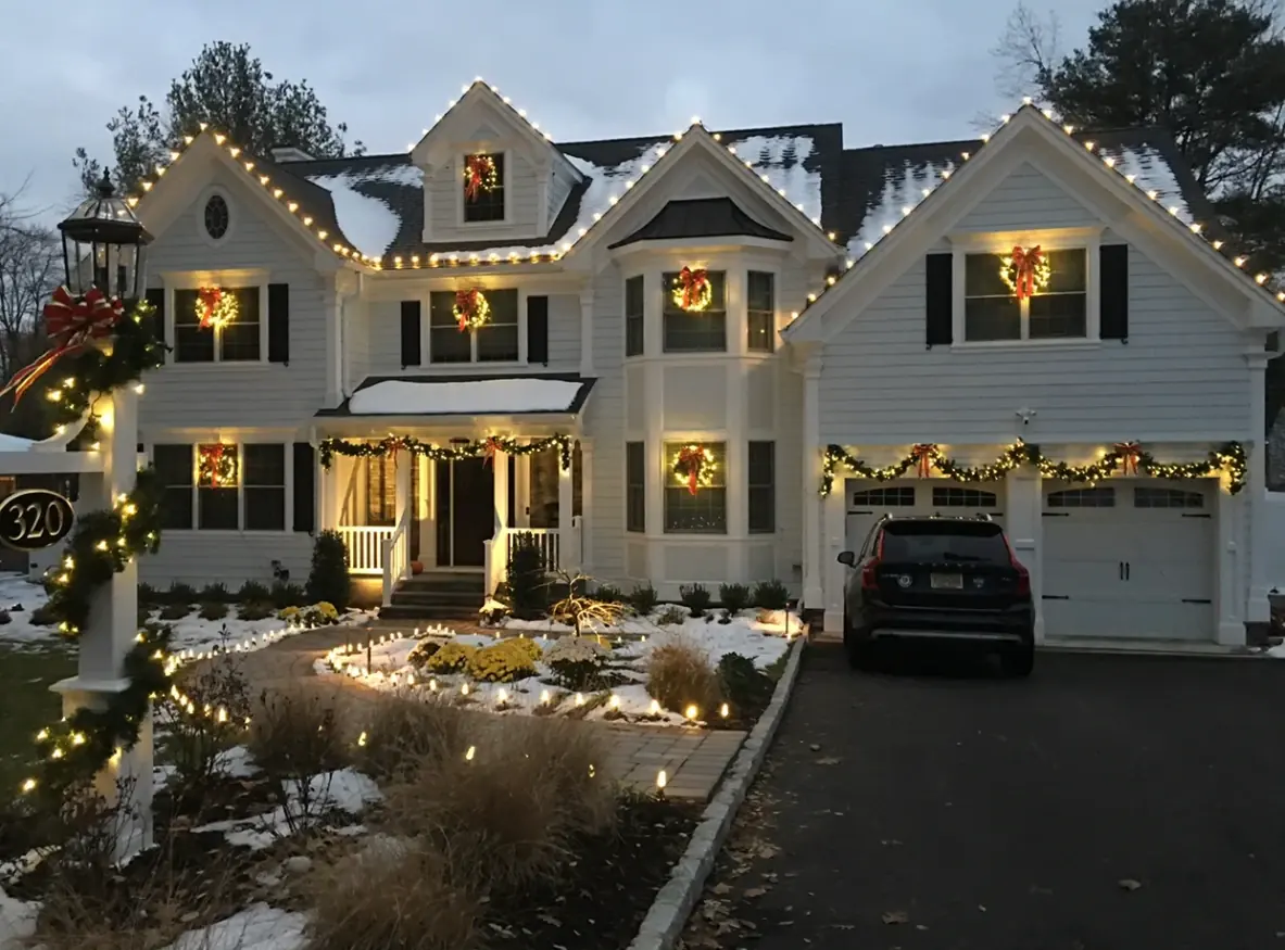 Snowy evening home with warm white roofline lights, lit window wreaths, garage garland and walkway lights