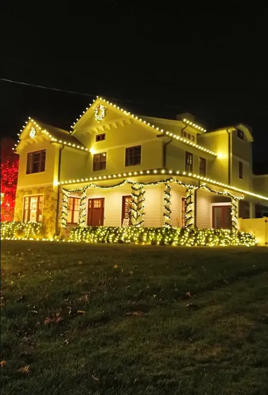 Two-story home with C9 roofline lights, garland-wrapped porch columns and lit hedges