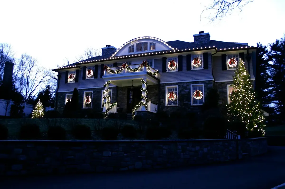Stone and shingle home at dusk with lit wreaths, spiral garland columns and a tall wrapped evergreen