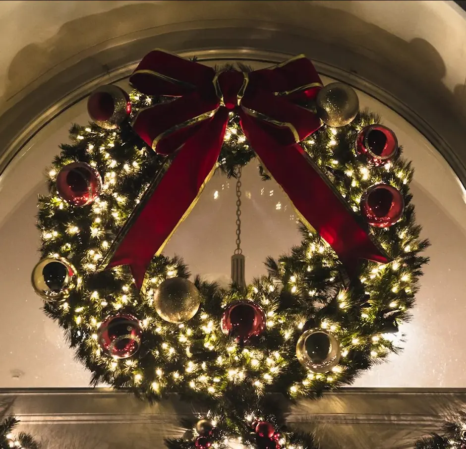 Large lit wreath with a velvet red bow and ornaments hung in an arched window